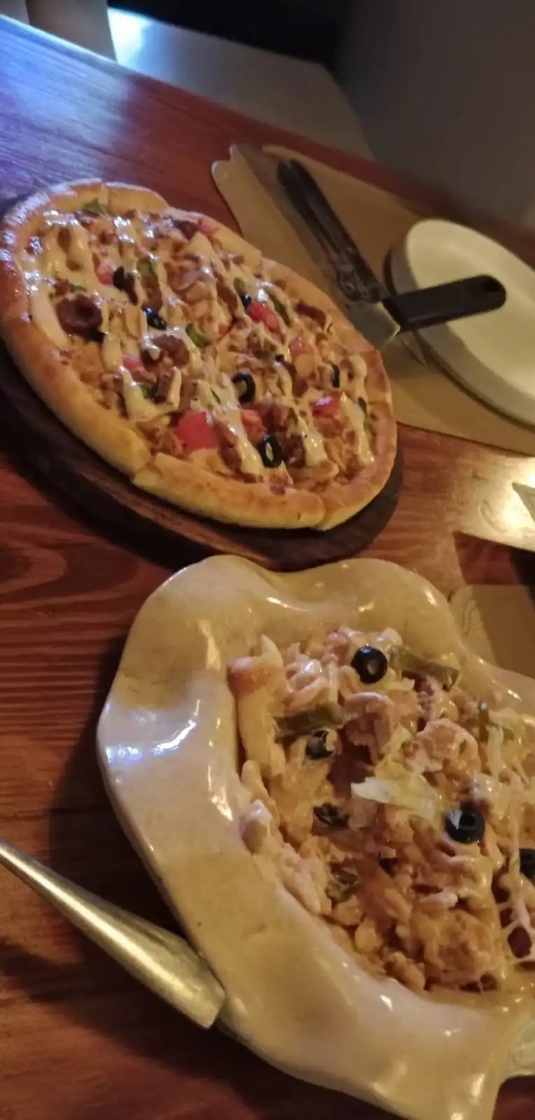 Two KAV pizzas on a wooden table with white plates and serving utensils, topped with cheese, meat and vegetables