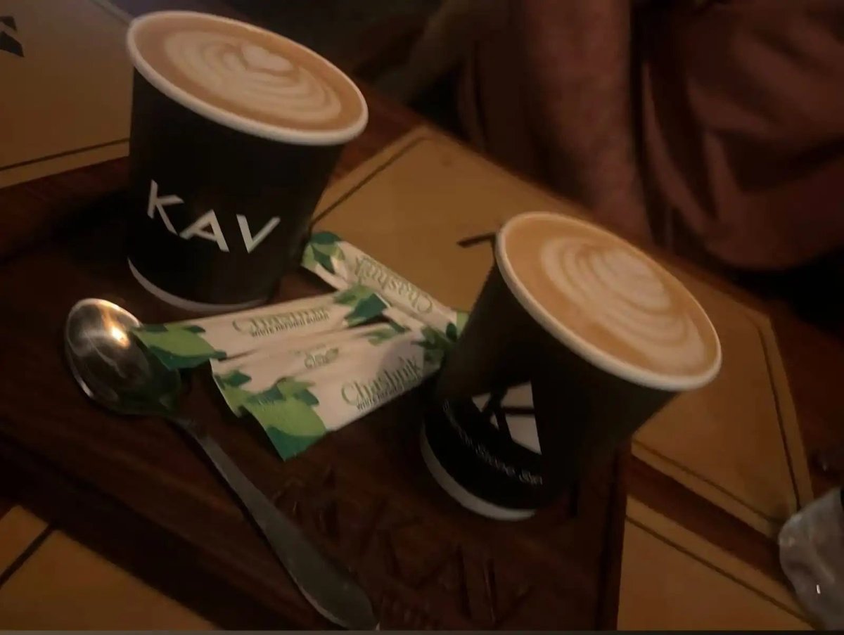 Two black KAV paper cups of latte with heart latte art, sugar packets and a spoon on a wooden tray