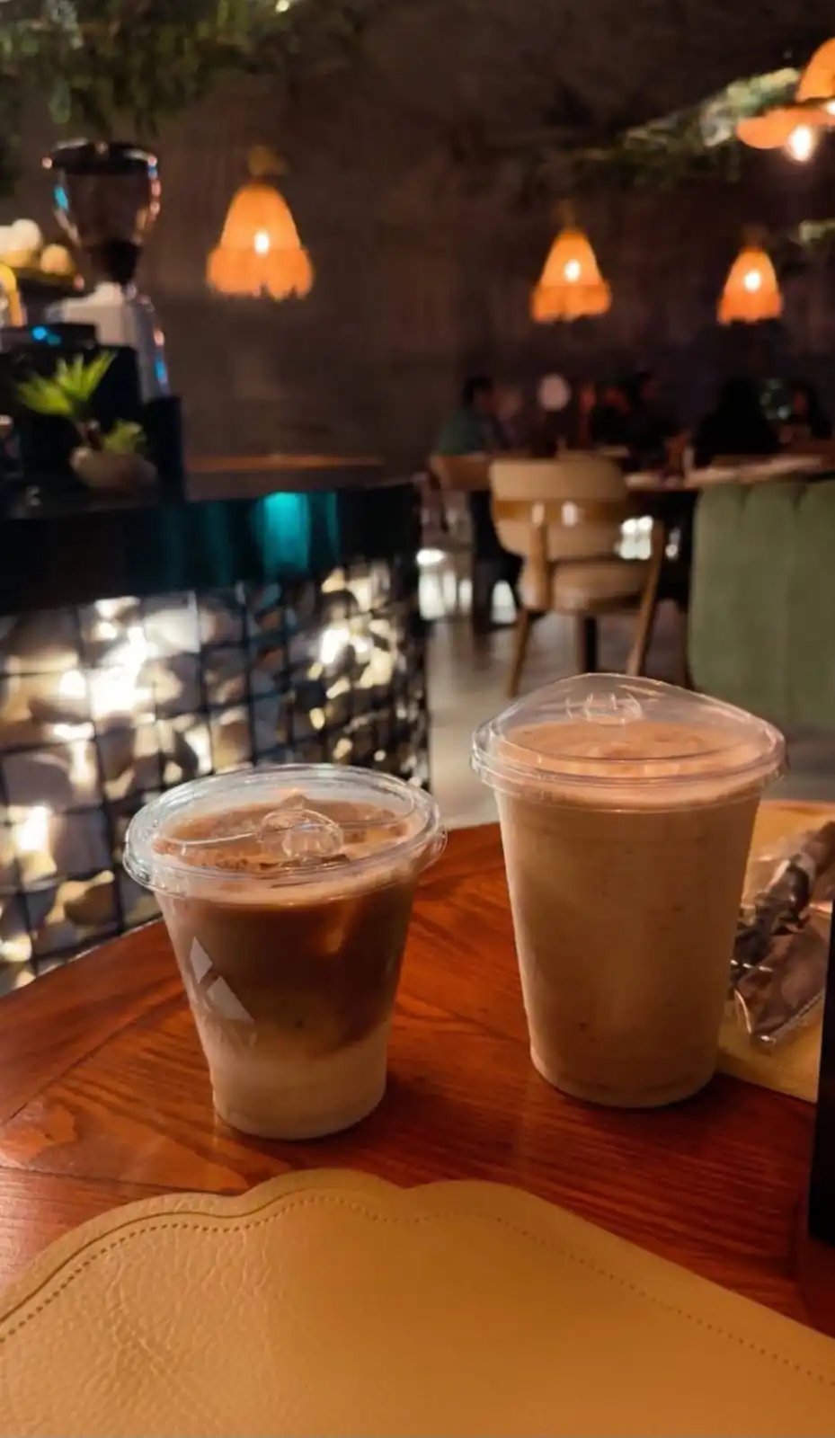 Two iced coffee drinks in KAV cups on a wooden table at a cozy café with warm orange pendant lights in the background