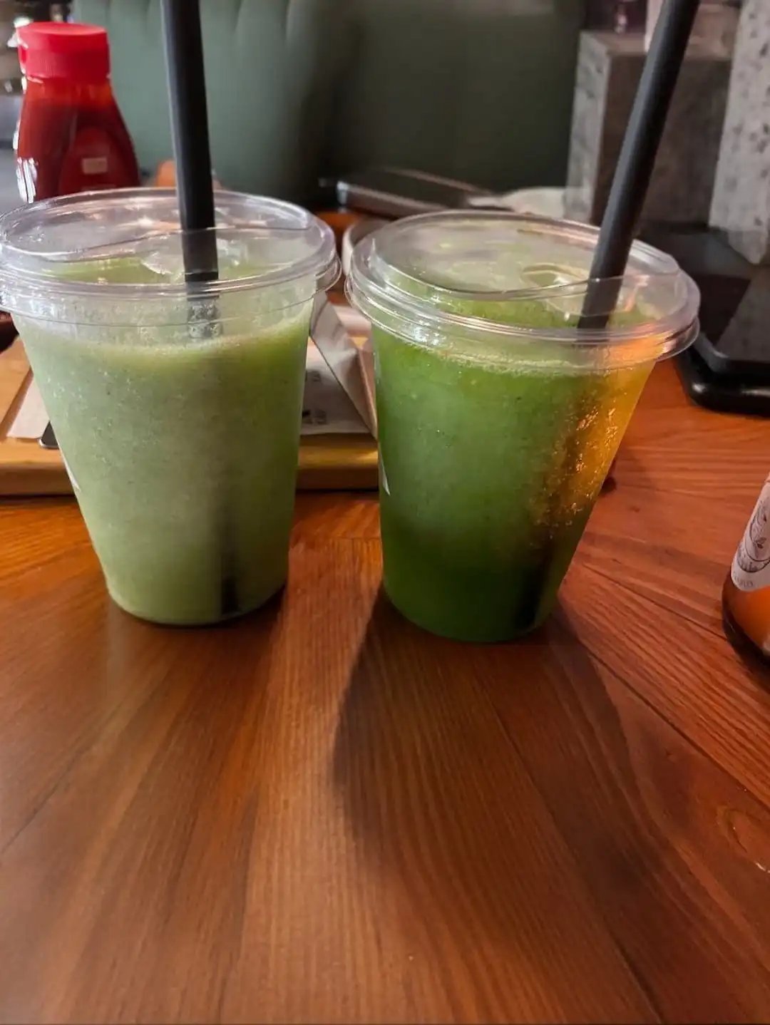 Two green juice drinks in clear plastic cups with black straws on a wooden café table