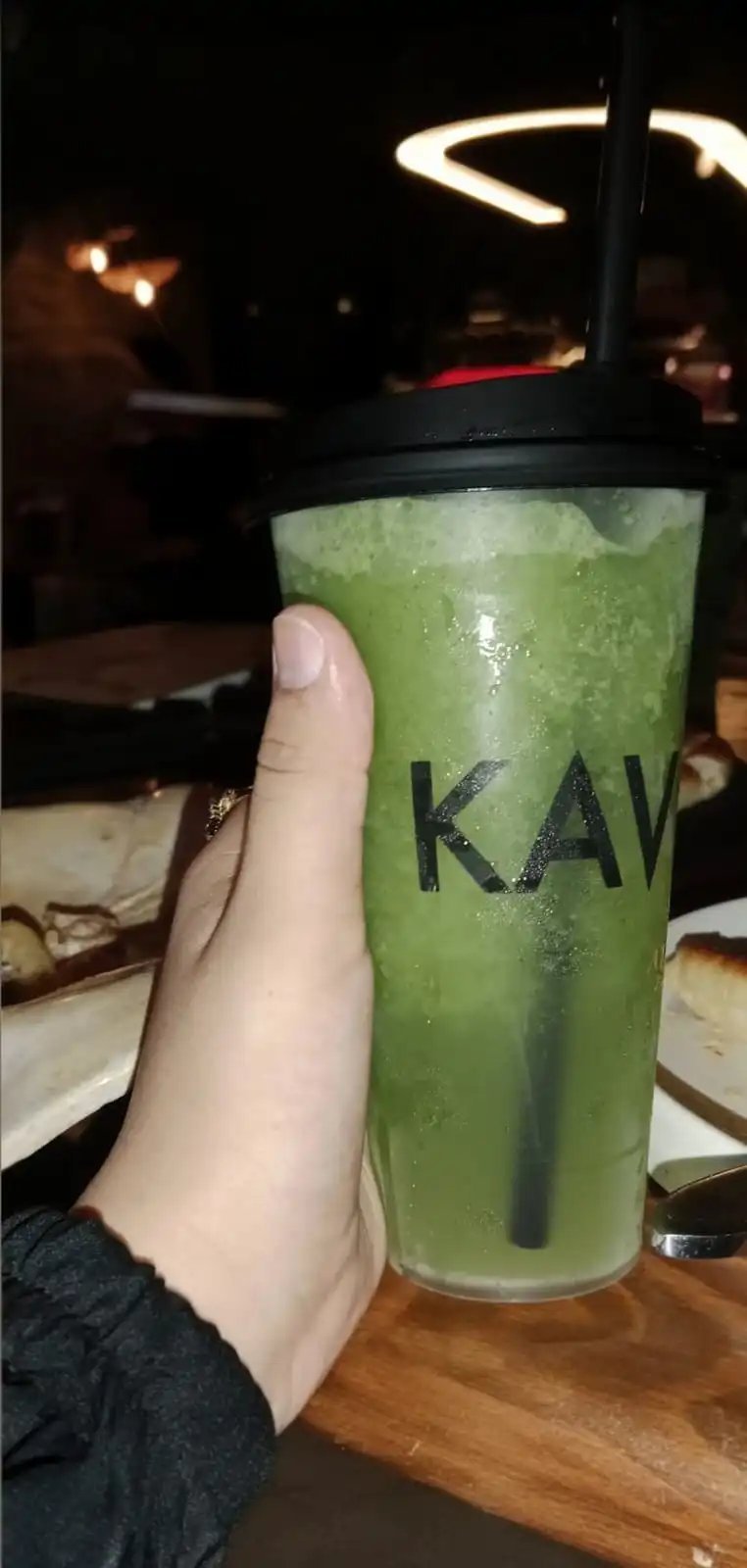 Hand holding a KAV branded green matcha drink in a clear cup with a black lid and straw at a dimly lit restaurant