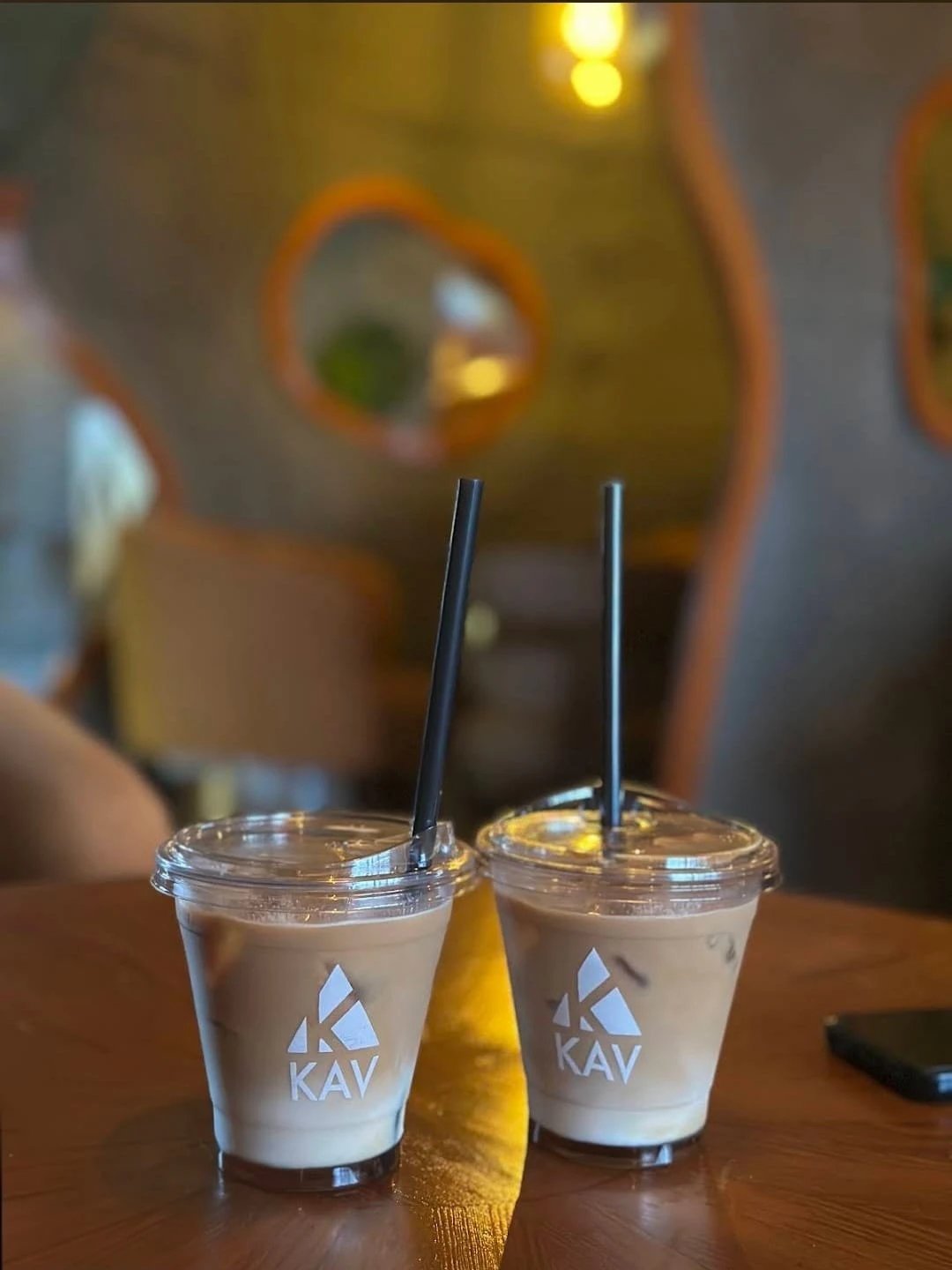 Two KAV branded iced beverages in clear plastic cups with black straws on a wooden table in a warm interior