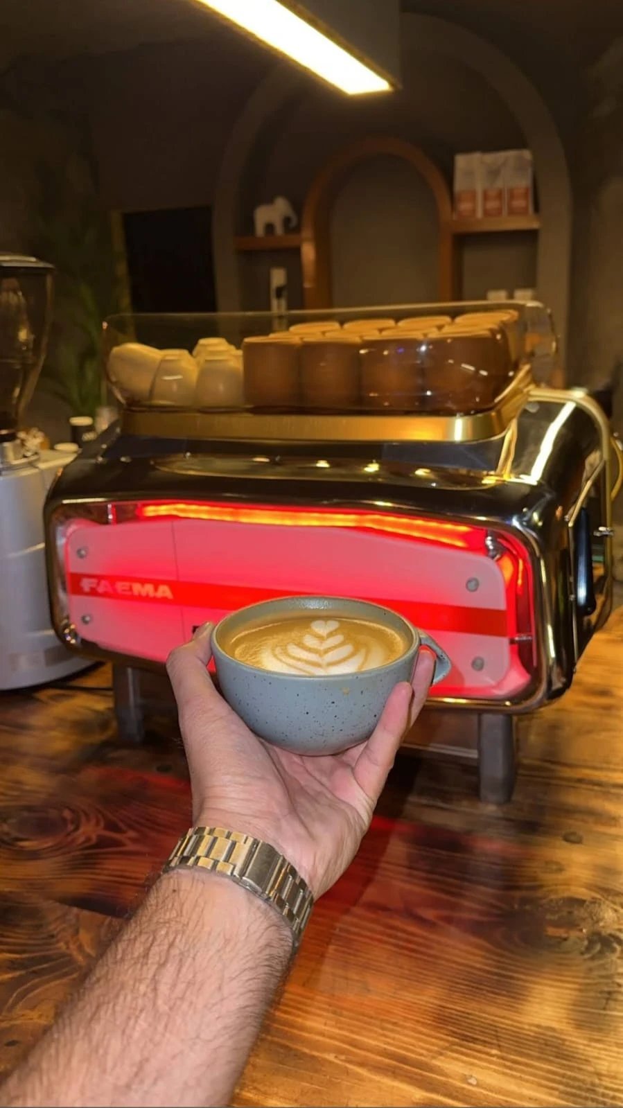 Hand holding a KAV latte in a grey speckled cup with leaf latte art, in front of a red Faema espresso machine