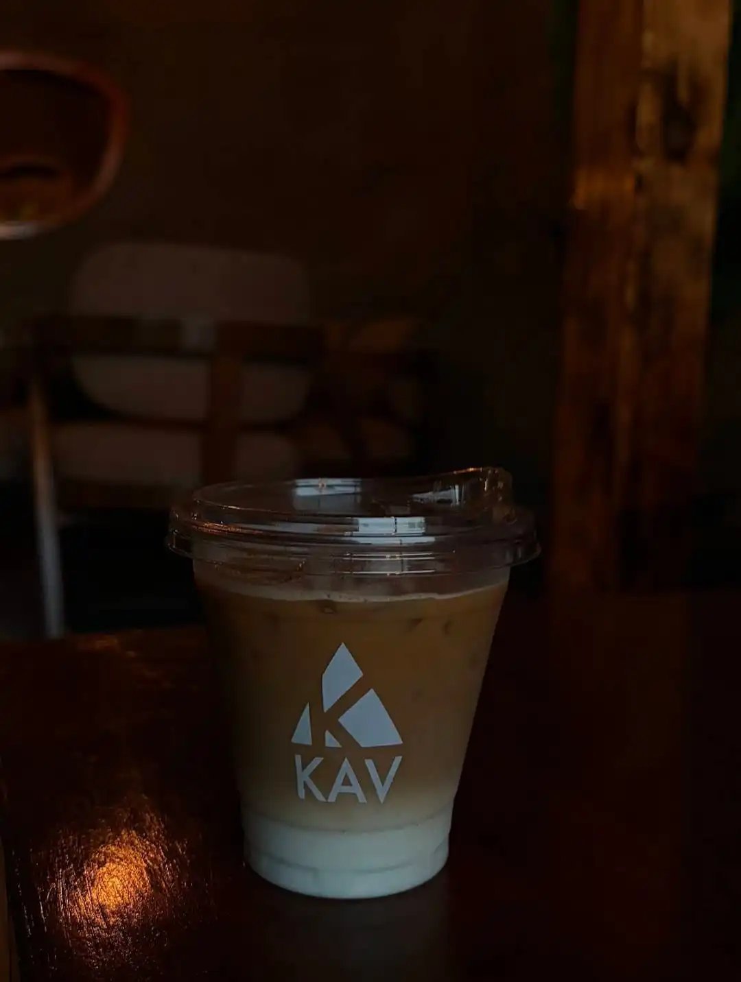 KAV branded iced coffee in a clear plastic cup with a white milk layer at the bottom, served on a dark surface in dim lighting