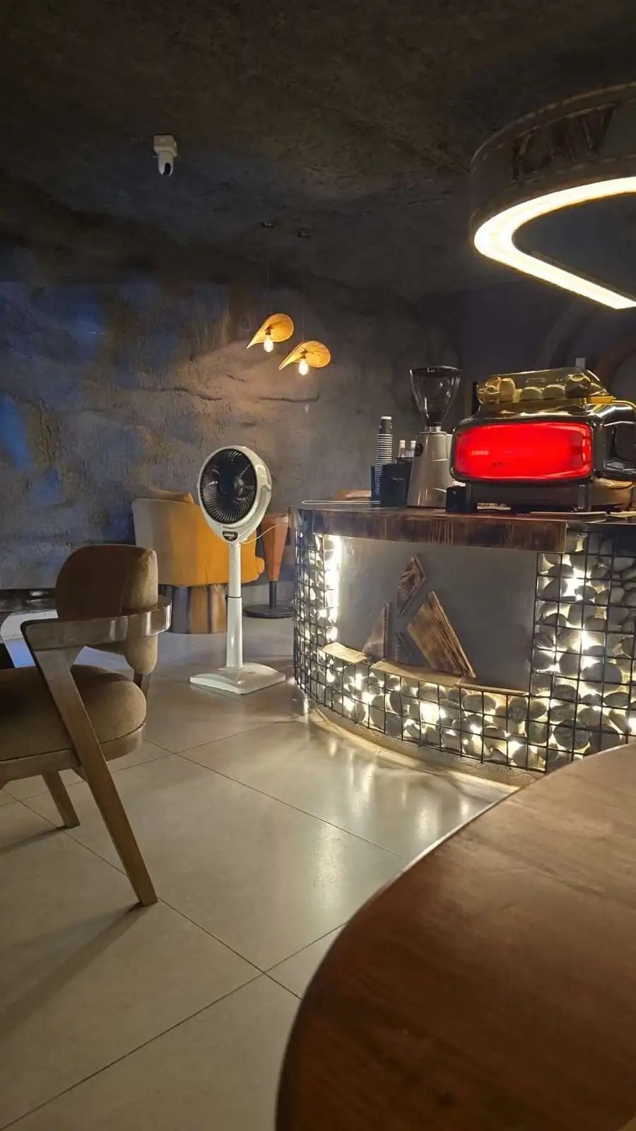 KAV coffee counter with the red espresso machine, stone-clad bar with a gabion shelf, and contemporary furniture in the cave-style dining room
