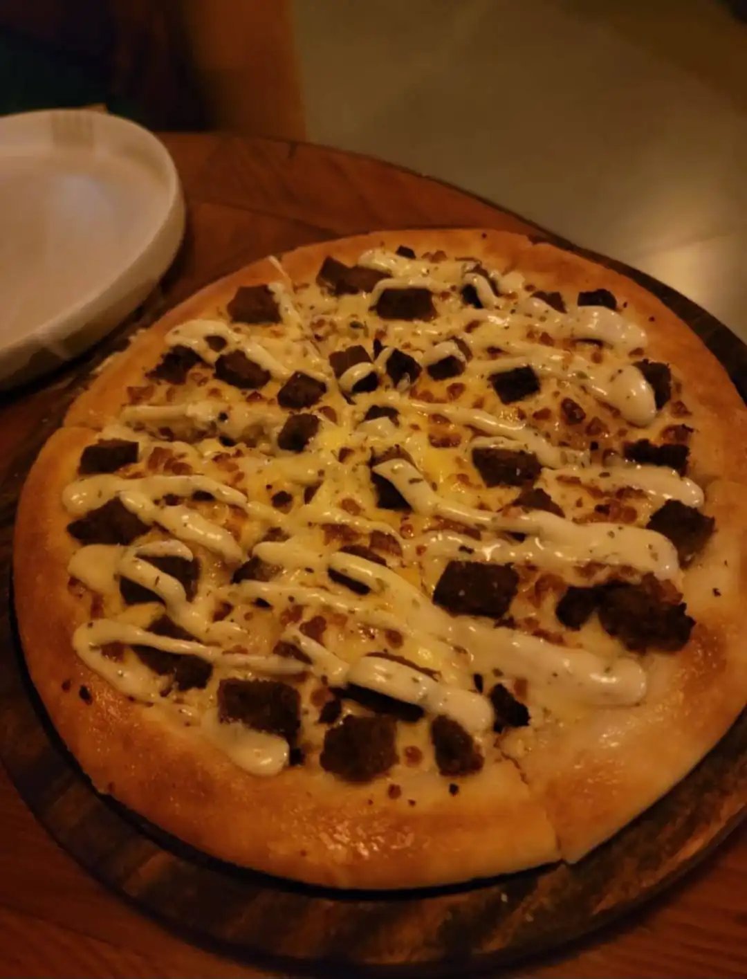 KAV pizza with white sauce, mozzarella and dark toppings drizzled on a wooden surface with a small white plate